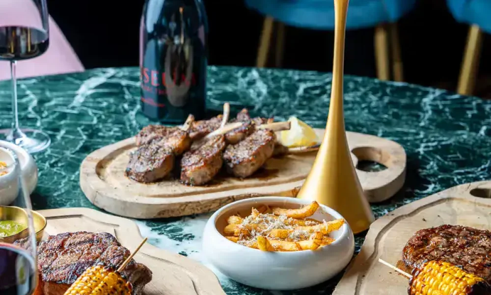 DSC01007 Gegrilltes Fleisch mit Maiskolben auf Holztellern, begleitet von Wein und cremiger Suppe, auf einem eleganten grünen Marmor-Tisch in einem modernen Restaurant.